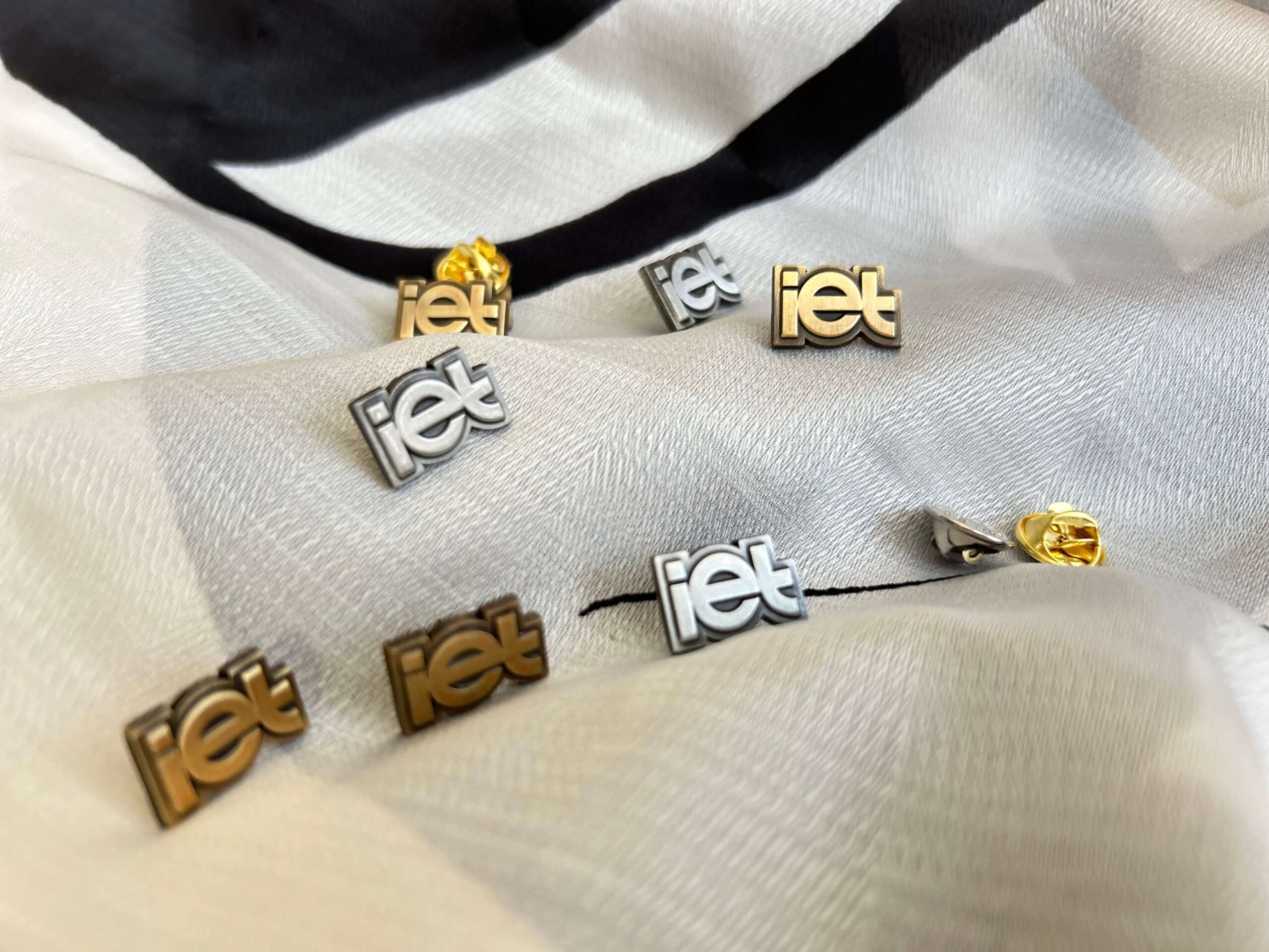 Zestaw personalizowanych przypinek firmowych z logo IET w kolorze złotym i srebrnym, ułożonych na eleganckim materiale.